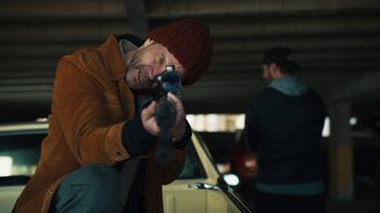 Movie still from “Sniper: Rogue Mission” (2022), directed by Oliver Thompson – A man holding a gun while standing next to a car; Medium shot, Low angle