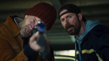 Movie still from “Sniper: Rogue Mission” (2022), directed by Oliver Thompson – Two men are looking at the camera while holding a gun; Close Up shot, Low angle