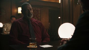 Movie still from “Sniper: Rogue Mission” (2022), directed by Oliver Thompson – An older man sitting at a table in front of a light; Medium shot, Over the shoulder angle