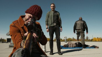 Movie still from “Sniper: Rogue Mission” (2022), directed by Oliver Thompson – A man standing next to another man holding a rifle; Wide shot, Low angle