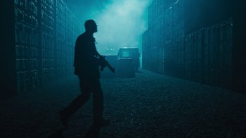 Movie still from “Sniper: Rogue Mission” (2022), directed by Oliver Thompson – A man is walking in the dark holding a gun; Wide shot, Low angle