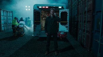 Movie still from “Sniper: Rogue Mission” (2022), directed by Oliver Thompson – A man standing next to an ambulance in the dark; Wide shot, Low angle
