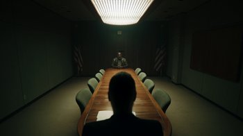 Movie still from “Sniper: Rogue Mission” (2022), directed by Oliver Thompson – A man sitting in front of a long table in front of american flags; Wide shot, Over the shoulder angle