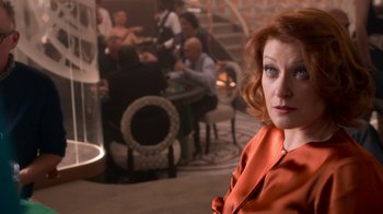 Movie still from “Snowpiercer” (2020), created by Graeme Manson – A woman in an orange dress in front of a group of people in a room; Medium shot, Over the shoulder angle
