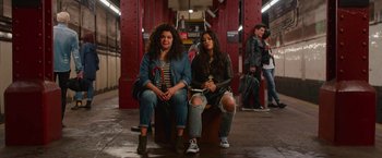 Movie still from “Someone Great” (2019), directed by Jennifer Kaytin Robinson – Two young women sitting on top of a bench in a train station; Wide shot, High angle