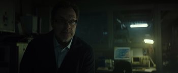 Movie still from “Spectral” (2016), directed by Nic Mathieu – A man in a suit and glasses is sitting in a dark room; Close Up shot, Low angle
