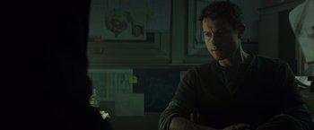 Movie still from “Spectral” (2016), directed by Nic Mathieu – A man sitting at a table in a dark room; Close Up shot, Over the shoulder angle