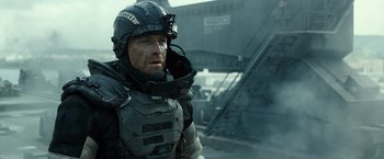Movie still from “Spectral” (2016), directed by Nic Mathieu – A man wearing a helmet and a body armor is standing outside; Medium shot, Over the shoulder angle