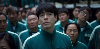 Movie still from “Squid Game” (2021), created by Hwang Dong-hyuk – A group of people standing around a man wearing a green jacket; Close Up shot, Low angle