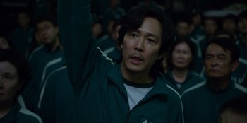 Movie still from “Squid Game” (2021), created by Hwang Dong-hyuk – A man in a green jacket is holding his hand up; Close Up shot, Low angle