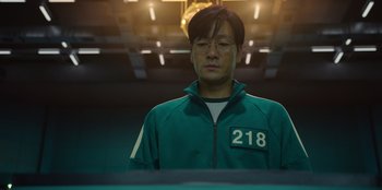 Movie still from “Squid Game” (2021), created by Hwang Dong-hyuk – A man wearing a green jacket standing in front of a table; Close Up shot, Low angle