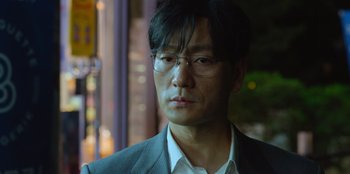 Movie still from “Squid Game” (2021), created by Hwang Dong-hyuk – A person wearing a suit and glasses; Close Up shot, Over the shoulder angle