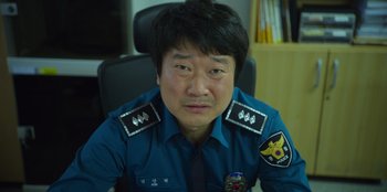 Movie still from “Squid Game” (2021), created by Hwang Dong-hyuk – A man in a police uniform sitting at a desk; Close Up shot, Low angle