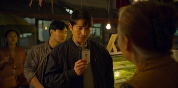 Movie still from “Squid Game” (2021), created by Hwang Dong-hyuk – A man holding up a cell phone in front of another man; Medium shot, Over the shoulder angle