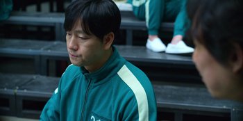 Movie still from “Squid Game” (2021), created by Hwang Dong-hyuk – A man sitting on some steps wearing a green jacket; Close Up shot, Over the shoulder angle