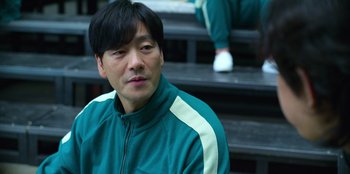 Movie still from “Squid Game” (2021), created by Hwang Dong-hyuk – A man wearing a green jacket sitting on some steps; Close Up shot, Over the shoulder angle