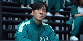 Movie still from “Squid Game” (2021), created by Hwang Dong-hyuk – A man sitting in front of a group of people; Close Up shot, Low angle