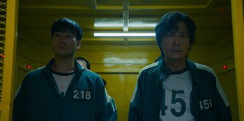 Movie still from “Squid Game” (2021), created by Hwang Dong-hyuk – Two asian men standing next to each other in front of a yellow wall; Medium shot, Low angle