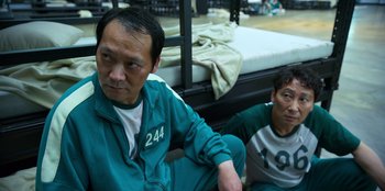 Movie still from “Squid Game” (2021), created by Hwang Dong-hyuk – Two men sitting next to each other in front of a bed; Medium shot, High angle