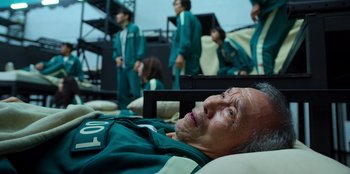 Movie still from “Squid Game” (2021), created by Hwang Dong-hyuk – An older man laying on a bed with a group of people in the background; Medium shot, High angle