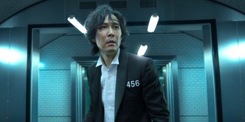 Movie still from “Squid Game” (2021), created by Hwang Dong-hyuk – A man in a black suit standing next to a door way; Close Up shot, Low angle