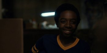 Movie still from “Stranger Things” (2016), created by Ross Duffer – A young man smiling for the camera in a dark room; Close Up shot, Over the shoulder angle