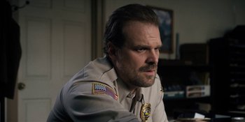 Movie still from “Stranger Things” (2016), created by Ross Duffer – A man in a uniform sitting in a room; Close Up shot, Over the shoulder angle