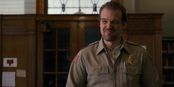 Movie still from “Stranger Things” (2016), created by Ross Duffer – A man in a uniform standing in front of a window; Close Up shot, Over the shoulder angle
