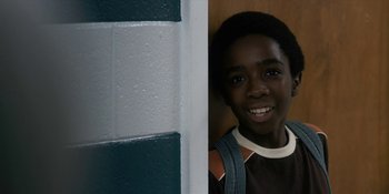 Movie still from “Stranger Things” (2016), created by Ross Duffer – A young man smiling at the camera next to a wall; Close Up shot, Over the shoulder angle