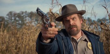 Movie still from “Stranger Things” (2016), created by Ross Duffer – A man holding a gun in a field; Medium shot, Low angle