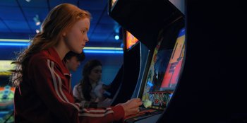 Movie still from “Stranger Things” (2016), created by Ross Duffer – A woman playing a video game in a game room; Medium shot, Over the shoulder angle