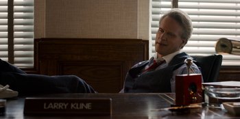 Movie still from “Stranger Things” (2016), created by Ross Duffer – A man sitting at a desk in front of a wooden sign; Medium shot, Over the shoulder angle