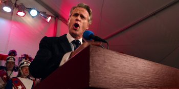 Movie still from “Stranger Things” (2016), created by Ross Duffer – A man in a suit is speaking into a microphone; Close Up shot, Low angle