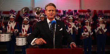 Movie still from “Stranger Things” (2016), created by Ross Duffer – A man in a suit and tie speaking at a podium; Medium shot, Low angle