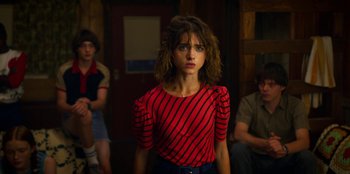 Movie still from “Stranger Things” (2016), created by Ross Duffer – A woman in a red and black striped shirt standing in front of a group of people; Medium shot, Over the shoulder angle