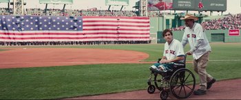 Movie still from “Stronger” (2017), directed by David Gordon Green – A man in a wheelchair on a baseball field with an american flag in the background; Wide shot, Over the shoulder angle