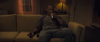 Movie still from “Synchronic” (2019), directed by Justin Benson – A man sitting on a couch drinking a beer while holding a remote; Medium shot, High angle