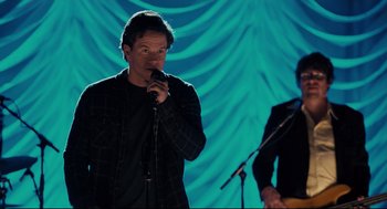 Movie still from “Ted” (2012), directed by Seth MacFarlane – A man holding a microphone while standing next to another man in front of a microphone stand; Medium shot, Over the shoulder angle