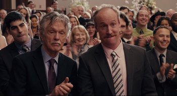 Movie still from “Ted” (2012), directed by Seth MacFarlane – A couple of men standing next to each other in front of a crowd; Medium shot, Over the shoulder angle
