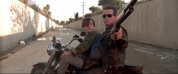 Movie still from “Terminator 2: Judgment Day” (1991), directed by James Cameron – A man and a woman riding a motorcycle down a street; Medium shot, Low angle