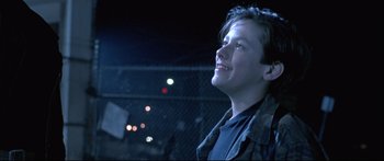 Movie still from “Terminator 2: Judgment Day” (1991), directed by James Cameron – A young man smiling while standing next to a chain link fence at night; Close Up shot, Low angle