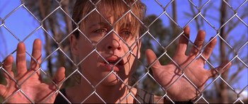 Movie still from “Terminator 2: Judgment Day” (1991), directed by James Cameron – A young man sticking out his tongue behind a chain link fence; Close Up shot, Low angle