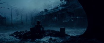 Movie still from “Terminator Genisys” (2015), directed by Alan Taylor – A person sitting on the ground in front of an abandoned house; Wide shot, Low angle