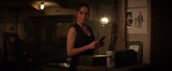 Movie still from “Terminator Genisys” (2015), directed by Alan Taylor – A woman holding a cell phone while standing in a room; Medium shot, Over the shoulder angle