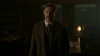 Movie still from “The Alienist” (2018), created by Cary Joji Fukunaga – A man in a suit and tie standing in a dark room; Medium shot, Low angle