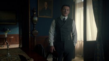 Movie still from “The Alienist” (2018), created by Cary Joji Fukunaga – A man standing in a room holding a chair; Medium shot, Over the shoulder angle