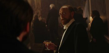 Movie still from “The Alienist” (2018), created by Cary Joji Fukunaga – A man in a tuxedo holding a glass of wine; Medium shot, Over the shoulder angle
