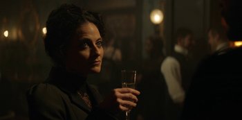 Movie still from “The Alienist” (2018), created by Cary Joji Fukunaga – A woman holding a glass of wine in a dark room; Close Up shot, Over the shoulder angle