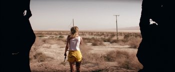 Movie still from “The Bad Batch” (2016), directed by Ana Lily Amirpour – A woman holding a gun while standing in the desert; Wide shot, Over the shoulder angle