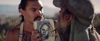 Movie still from “The Bad Batch” (2016), directed by Ana Lily Amirpour – A man holding up a drawing of a woman to a man; Close Up shot, Over the shoulder angle